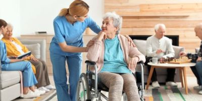 Older people in a care home setting. Two are playing chess and one person is looking at a book with a carer. Another person is sitting in a wheelchair talking to a carer.