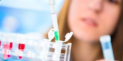 Close up of a green liquid being transferred from a pipette into a small test tube - with a blurred scientist headshot in the background.