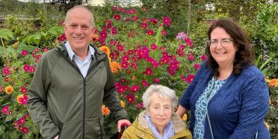 Family photo of Rosie Woollard and her two children, Bernard Woollard (left) and Sue Pavitt (right). They are smiling in front of a backdrop of flowers.