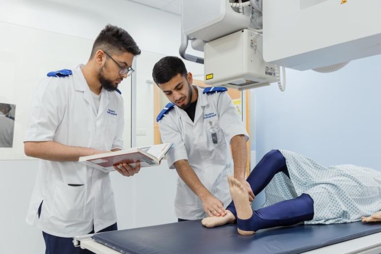 Two male students in clinical attire, one interacting with a dummy's feet laid on a table, the other consulting a radiography textbook.